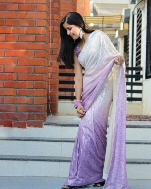Woman in a shimmering purple saree stands gracefully, showcasing traditional Indian fashion.