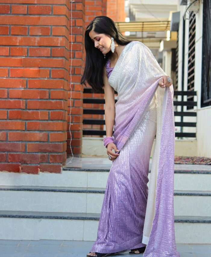 Woman in a shimmering purple saree stands gracefully, showcasing traditional Indian fashion.