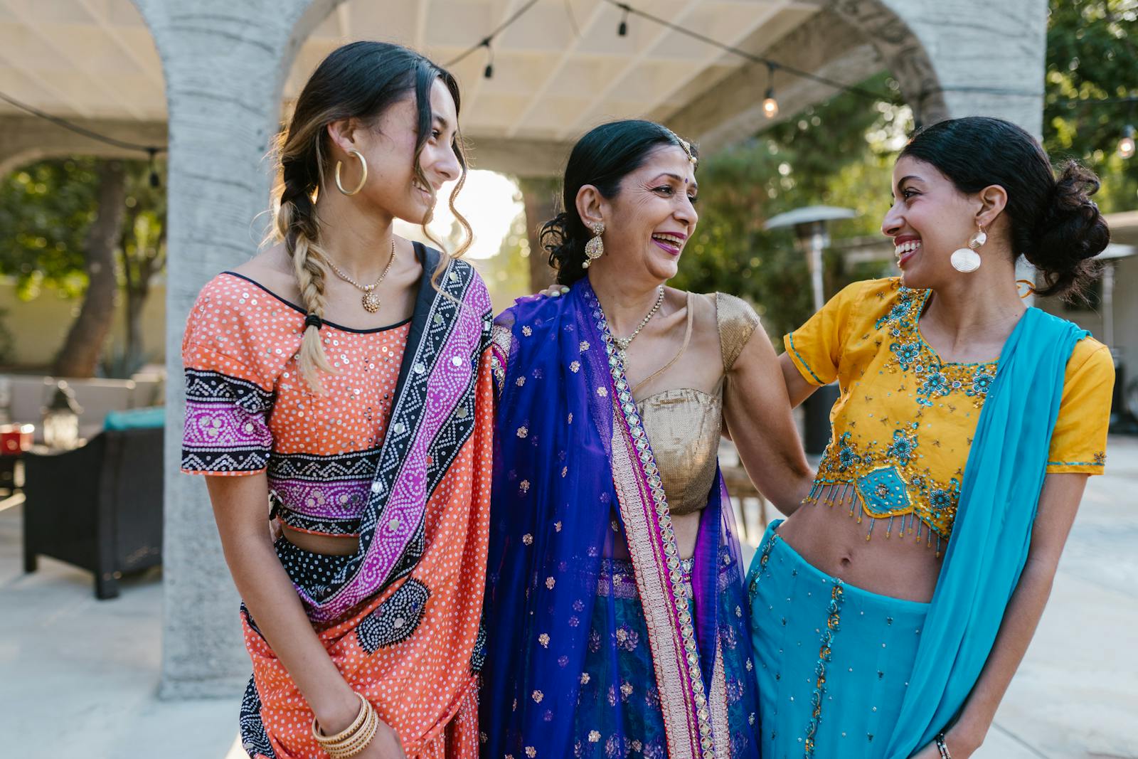 Three women smiling and embracing in colorful Indian saris, celebrating a joyous occasion outdoors.